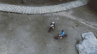 An aerial view of two cyclists wearing helmets and biking gear, positioned on a paved path surrounded by a mix of grass and dirt terrain. One cyclist wears a blue jacket, while the other is in black. A large rock formation is visible in the bottom right corner.