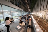 A group of people are seated at tables arranged in a linear fashion along a corridor. The setting appears to be a modern, indoor space with glass walls on one side and a series of bamboo plants on the other. There is a woman standing and speaking, possibly addressing the group. The scene is illuminated by overhead lights and string lights along the bamboo side.