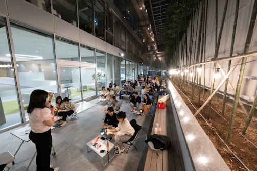 A group of people are seated at tables arranged in a linear fashion along a corridor. The setting appears to be a modern, indoor space with glass walls on one side and a series of bamboo plants on the other. There is a woman standing and speaking, possibly addressing the group. The scene is illuminated by overhead lights and string lights along the bamboo side.