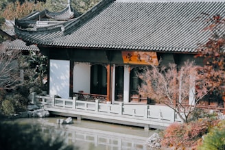 A traditional Asian-style building with a sweeping tiled roof and wooden pillars. The architecture is surrounded by lush greenery and a small pond, creating a serene and peaceful atmosphere.