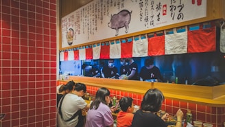 People are sitting at a counter in a ramen restaurant, eating and facing a kitchen area where chefs are preparing food. Red and white tiles cover the walls, and above the counter, there is a decorative cloth with a pattern that matches. A banner with Japanese text and a pig illustration is displayed prominently.