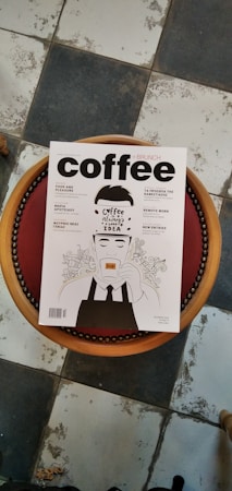 A magazine with the title 'coffee + brunch' is placed on a round, cushioned stool with a wooden edge. The cover features a stylized illustration of a person holding a cup of coffee, surrounded by artistic line drawings. The floor beneath is a worn, black and white tile pattern.