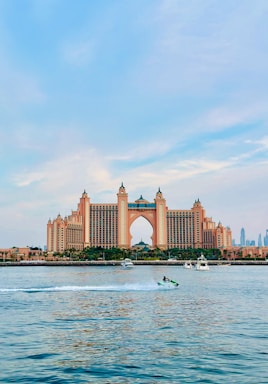 A large, luxurious hotel with a central archway and multiple towers stands prominently on the waterfront. The architecture is grand and symmetrical, featuring peach and pink tones. In front of the hotel, a body of water reflects the building, showing several boats, including a person riding a jet ski over the water's surface. The sky above is bright with light clouds, adding a calm atmosphere to the scene.