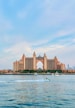 A large, luxurious hotel with a central archway and multiple towers stands prominently on the waterfront. The architecture is grand and symmetrical, featuring peach and pink tones. In front of the hotel, a body of water reflects the building, showing several boats, including a person riding a jet ski over the water's surface. The sky above is bright with light clouds, adding a calm atmosphere to the scene.