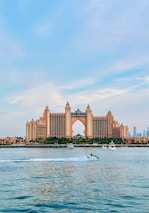 A large, luxurious hotel with a central archway and multiple towers stands prominently on the waterfront. The architecture is grand and symmetrical, featuring peach and pink tones. In front of the hotel, a body of water reflects the building, showing several boats, including a person riding a jet ski over the water's surface. The sky above is bright with light clouds, adding a calm atmosphere to the scene.