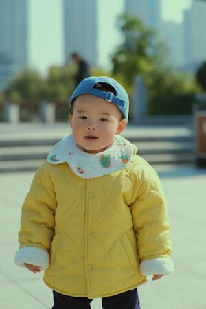 A young child wearing a vibrant yellow quilted jacket and a blue cap standing in an outdoor setting. The backdrop includes blurred greenery and urban architecture, indicating a city park or plaza.