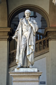A marble statue of a man in formal attire, depicted in a thoughtful pose with one hand touching his chin, stands on a pedestal. The figure is draped in a long coat and holds a scroll in his other hand. The sculpture is situated in front of an architectural backdrop featuring arches and stairs.