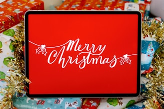 A digital display with the words 'Merry Christmas' written in elegant white script, surrounded by festive wrapping paper and decorative items. The display is set against a vibrant red background, with gold tinsel adding a touch of sparkle.