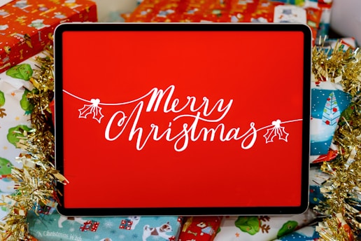 A digital display with the words 'Merry Christmas' written in elegant white script, surrounded by festive wrapping paper and decorative items. The display is set against a vibrant red background, with gold tinsel adding a touch of sparkle.