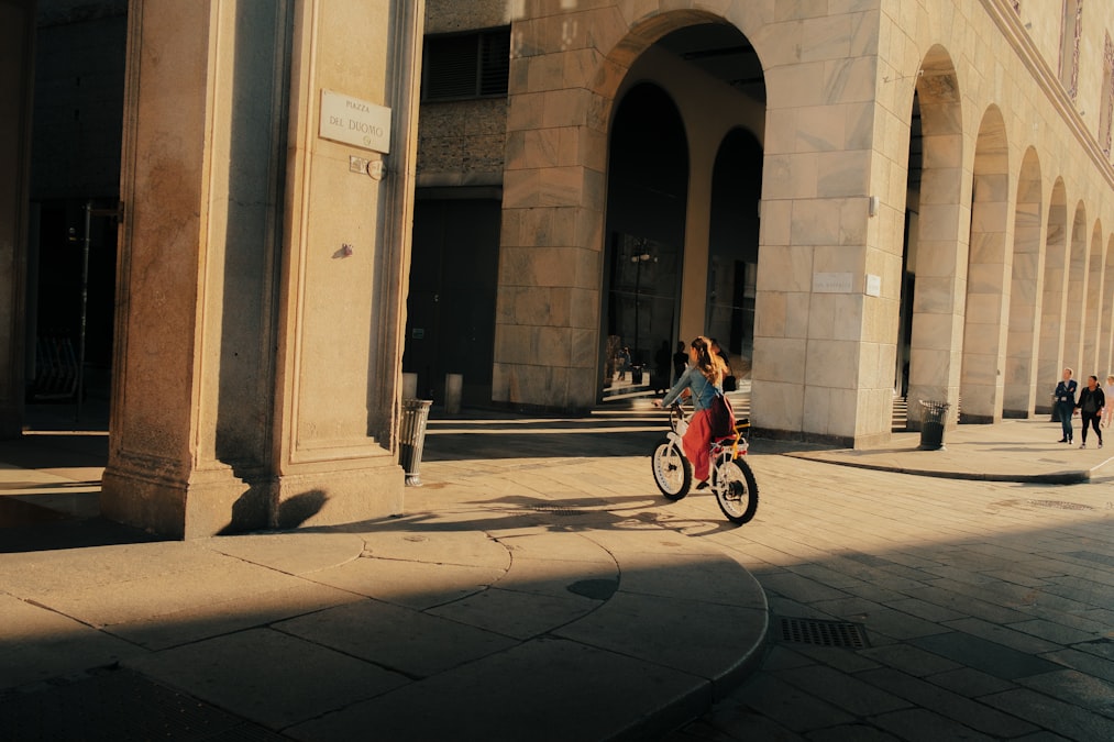 Mulher pedalando uma bicicleta por uma praça histórica de pedra ao entardecer na cidade