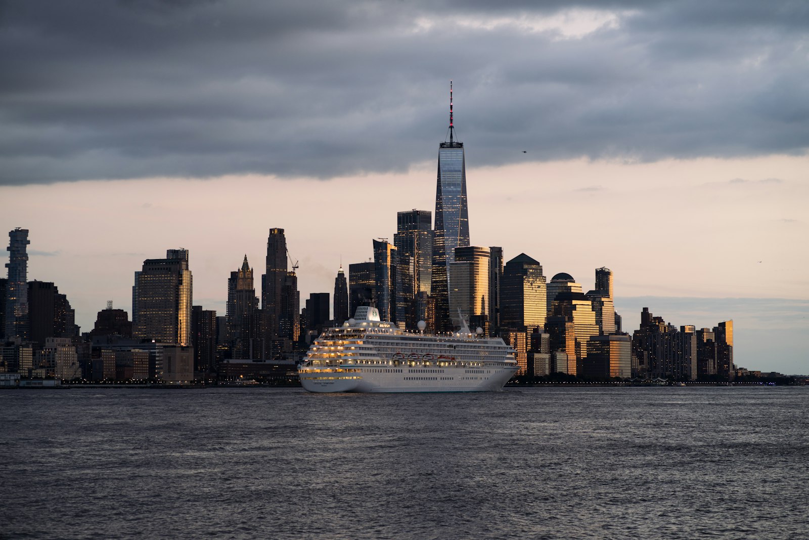 Cruise ship on the Hudson River in front of the Lower Manhattan skyline and One World Trade Center under a cloudy sunset sky