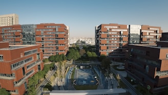 Tall, red brick buildings surround an open courtyard with pathways and a small sports field. Trees and greenery line the pathways, and there is a clear blue sky overhead.