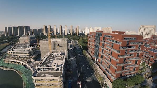 An urban landscape featuring modern high-rise buildings and a construction site. The foreground displays red brick buildings with an ongoing construction project on the left, including cranes and unfinished structures. A road divides the area, leading towards a background of more high-rise buildings under a clear blue sky.