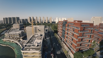 An urban landscape featuring modern high-rise buildings and a construction site. The foreground displays red brick buildings with an ongoing construction project on the left, including cranes and unfinished structures. A road divides the area, leading towards a background of more high-rise buildings under a clear blue sky.