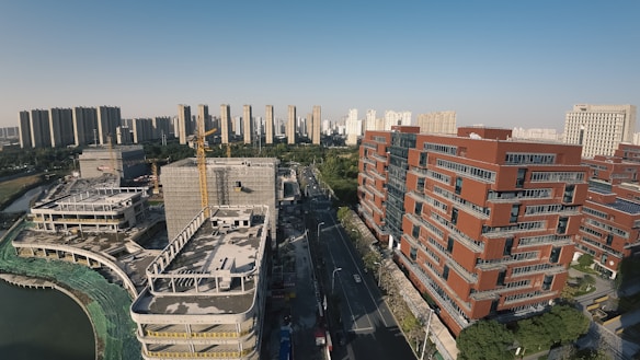 An urban landscape featuring modern high-rise buildings and a construction site. The foreground displays red brick buildings with an ongoing construction project on the left, including cranes and unfinished structures. A road divides the area, leading towards a background of more high-rise buildings under a clear blue sky.