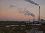 A large industrial facility with several smokestacks emits clouds of smoke against a sunset sky. The foreground features a densely populated area with rooftops and trees, while the industrial complex dominates the background.