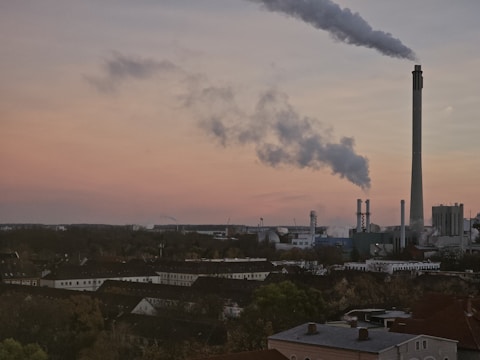 A large industrial facility with several smokestacks emits clouds of smoke against a sunset sky. The foreground features a densely populated area with rooftops and trees, while the industrial complex dominates the background.