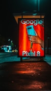 An illuminated advertising display at night features a smartphone held by a hand with a visible ring. The text on the display includes 'Google' and 'Pixel 8', indicating a product advertisement. The background is an urban setting with a street and scattered lights.