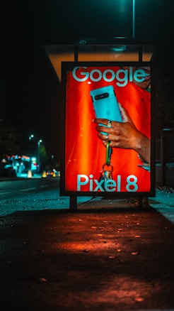 An illuminated advertising display at night features a smartphone held by a hand with a visible ring. The text on the display includes 'Google' and 'Pixel 8', indicating a product advertisement. The background is an urban setting with a street and scattered lights.