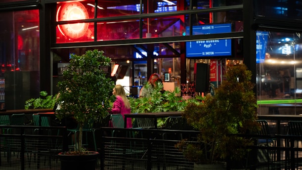 A dimly lit bar or restaurant interior with a modern design, featuring large windows, greenery, and neon lighting. Signs and screens are visible, indicating a casual and lively atmosphere. People are seated at tables, suggesting social interaction.