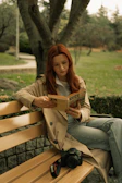 A candid shot of a woman sitting on a park bench in a light trench coat, reading a book under gentle afternoon light.
