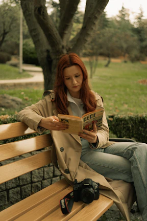 A candid shot of a woman sitting on a park bench in a light trench coat, reading a book under gentle afternoon light.