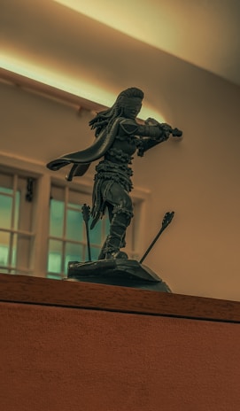 A sculpture of a warrior in an action pose stands on a ledge indoors. The warrior holds a weapon and is surrounded by arrows embedded in the ground, suggesting a dynamic and intense scene. The background features windows and an overhead light, casting a soft glow.
