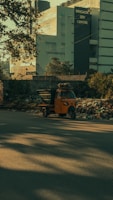 A bright orange auto rickshaw is parked by the side of a road, surrounded by a pile of rubbish and debris. In the background, there is a large modern building with the sign 'Brigade IRV Centre.' Trees and shadows are visible, adding to the overall urban environment.