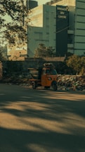 A bright orange auto rickshaw is parked by the side of a road, surrounded by a pile of rubbish and debris. In the background, there is a large modern building with the sign 'Brigade IRV Centre.' Trees and shadows are visible, adding to the overall urban environment.