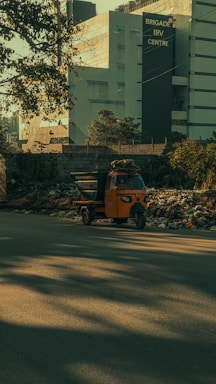 A bright orange auto rickshaw is parked by the side of a road, surrounded by a pile of rubbish and debris. In the background, there is a large modern building with the sign 'Brigade IRV Centre.' Trees and shadows are visible, adding to the overall urban environment.