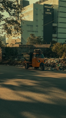 A bright orange auto rickshaw is parked by the side of a road, surrounded by a pile of rubbish and debris. In the background, there is a large modern building with the sign 'Brigade IRV Centre.' Trees and shadows are visible, adding to the overall urban environment.