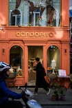 A brightly colored storefront for a kids and baby clothing shop with a sign reading 'bondberro.' The window displays various children's clothing items, and a person in dark clothing is entering the shop. In the foreground, a person on a scooter is passing by.