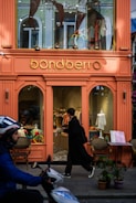 A brightly colored storefront for a kids and baby clothing shop with a sign reading 'bondberro.' The window displays various children's clothing items, and a person in dark clothing is entering the shop. In the foreground, a person on a scooter is passing by.
