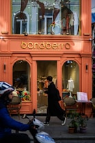 A brightly colored storefront for a kids and baby clothing shop with a sign reading 'bondberro.' The window displays various children's clothing items, and a person in dark clothing is entering the shop. In the foreground, a person on a scooter is passing by.
