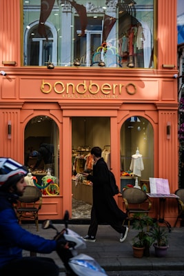 A brightly colored storefront for a kids and baby clothing shop with a sign reading 'bondberro.' The window displays various children's clothing items, and a person in dark clothing is entering the shop. In the foreground, a person on a scooter is passing by.