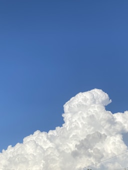 A large formation of fluffy white cumulus clouds rises against a clear blue sky. The clouds appear dense and textured, showcasing various shades of white and hinting at the soft, cotton-like quality.