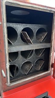 A metallic chamber with multiple circular openings arranged in two rows. Each opening has triangular metal plates inside. The exterior frame is bordered with a fibrous material, and the lower section displays a red surface.