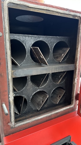 A metallic chamber with multiple circular openings arranged in two rows. Each opening has triangular metal plates inside. The exterior frame is bordered with a fibrous material, and the lower section displays a red surface.
