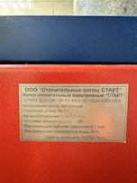 A close-up of a metal plaque with text in Russian, affixed to a red and blue industrial machine. The text provides technical specifications for a heating boiler, including model, serial number, power capacity, pressure, and temperature limits.