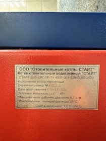 A close-up of a metal plaque with text in Russian, affixed to a red and blue industrial machine. The text provides technical specifications for a heating boiler, including model, serial number, power capacity, pressure, and temperature limits.