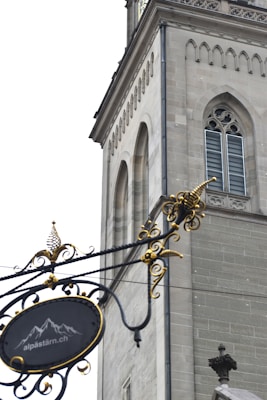 An ornate wrought iron sign hangs in the foreground, featuring decorative swirls and gold accents. In the background, a stone building with Gothic architectural elements, including arched windows and detailed masonry, is visible. The sign includes a motif of mountains and a website URL.