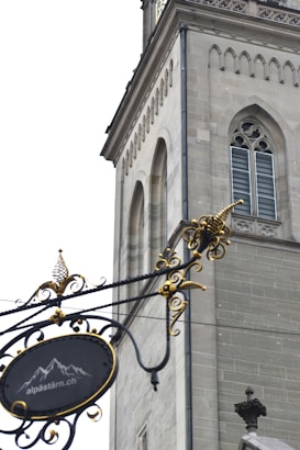 An ornate wrought iron sign hangs in the foreground, featuring decorative swirls and gold accents. In the background, a stone building with Gothic architectural elements, including arched windows and detailed masonry, is visible. The sign includes a motif of mountains and a website URL.