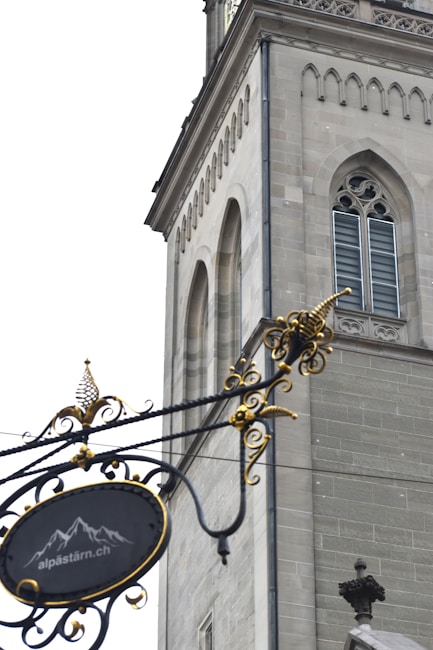 An ornate wrought iron sign hangs in the foreground, featuring decorative swirls and gold accents. In the background, a stone building with Gothic architectural elements, including arched windows and detailed masonry, is visible. The sign includes a motif of mountains and a website URL.