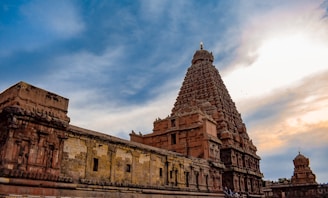 A large, intricately designed temple with a towering central structure made of stone. The sky above is partly cloudy, with hints of blue and orange from the setting or rising sun. The temple's architecture features detailed carvings and multiple levels, indicating its historical and cultural significance.