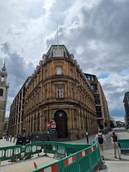 An ornate historic building with a dome roof dominates the street view, flanked by modern structures. The entrance reads 'City of London Magistrates Court', with a restricted access sign and construction barriers in the foreground. Pedestrians are walking near the building under a partly cloudy sky.