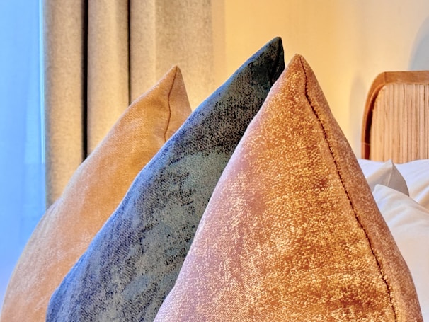 Three decorative pillows positioned in a bedroom setting. The pillows have a velvet-like texture and come in shades of gold, dark blue, and light tan. They are arranged in front of a partially visible wooden headboard. Pleated curtains are drawn in the background, and soft lighting creates a cozy atmosphere.