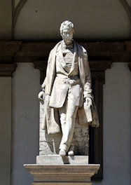 A marble statue depicting a man in a formal 19th-century style outfit, including a waistcoat and long coat, is positioned against a decorative stone background. The figure appears composed and dignified, with one leg slightly forward, suggesting movement or confidence. The base of the statue is inscribed with the name 'Tommaso Grossi.' The structure is set in an architectural niche with arches.