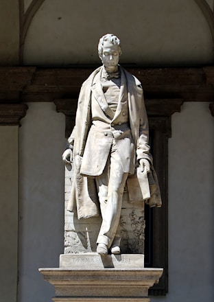 A marble statue depicting a man in a formal 19th-century style outfit, including a waistcoat and long coat, is positioned against a decorative stone background. The figure appears composed and dignified, with one leg slightly forward, suggesting movement or confidence. The base of the statue is inscribed with the name 'Tommaso Grossi.' The structure is set in an architectural niche with arches.
