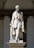 A marble statue depicting a man in a formal 19th-century style outfit, including a waistcoat and long coat, is positioned against a decorative stone background. The figure appears composed and dignified, with one leg slightly forward, suggesting movement or confidence. The base of the statue is inscribed with the name 'Tommaso Grossi.' The structure is set in an architectural niche with arches.