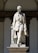 A marble statue depicting a man in a formal 19th-century style outfit, including a waistcoat and long coat, is positioned against a decorative stone background. The figure appears composed and dignified, with one leg slightly forward, suggesting movement or confidence. The base of the statue is inscribed with the name 'Tommaso Grossi.' The structure is set in an architectural niche with arches.